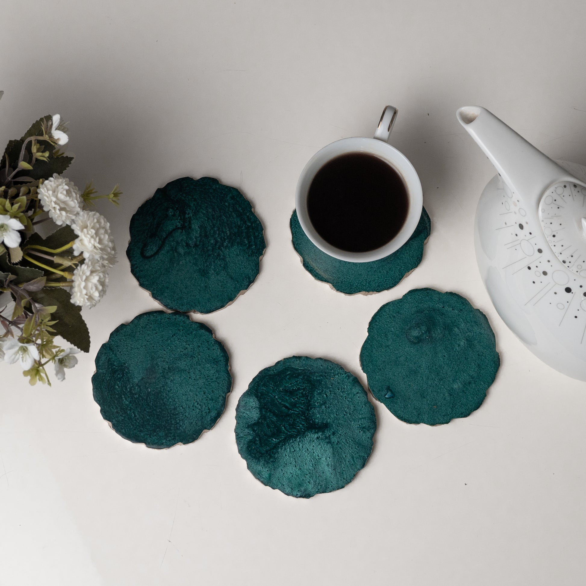 Teal coasters with a cup of coffee and teapot on a light surface.